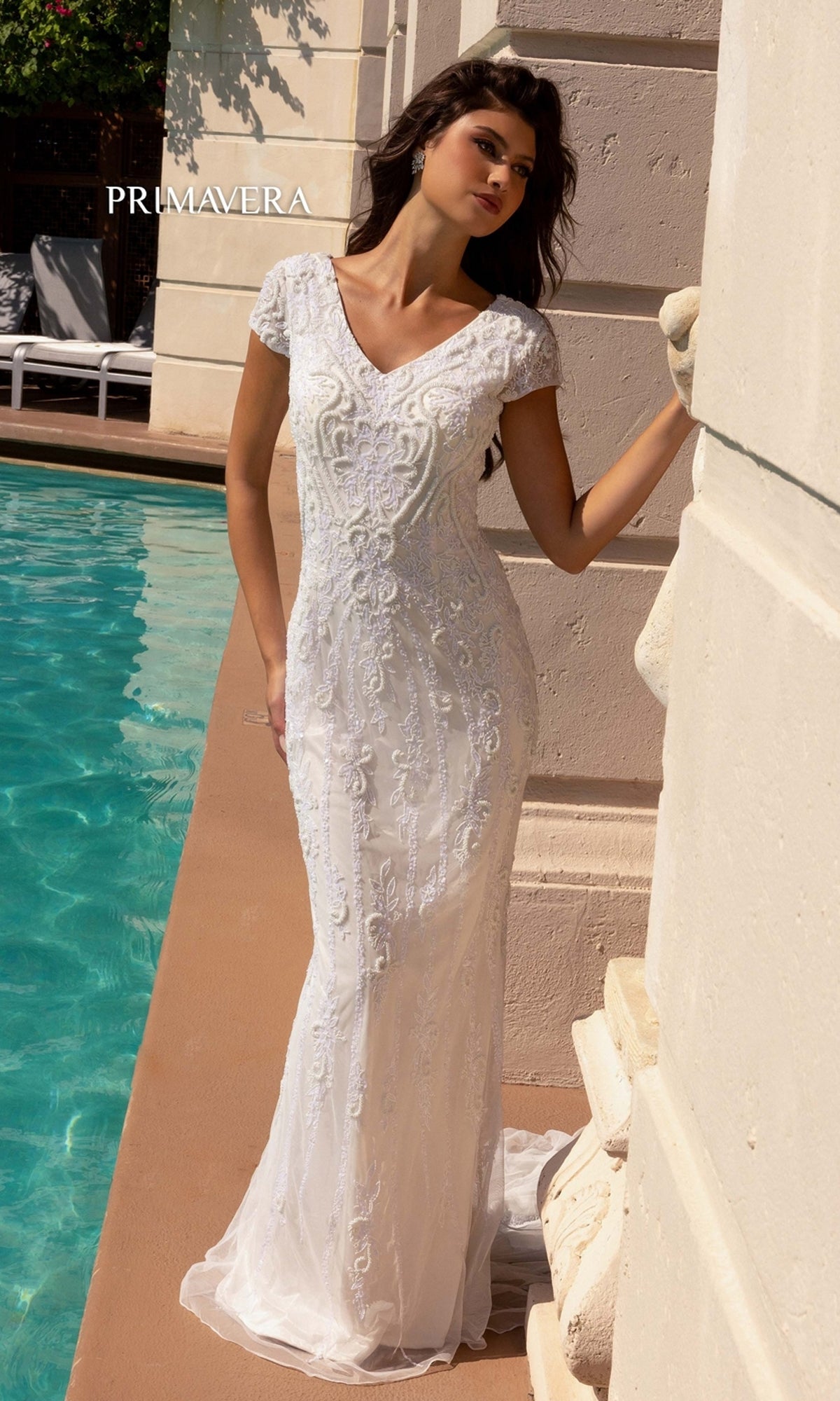 A woman in the Long Wedding Dress 11121 by Primavera—a white, embellished, short-sleeve gown with beaded embroidery and v-neckline—stands poolside against a light wall.